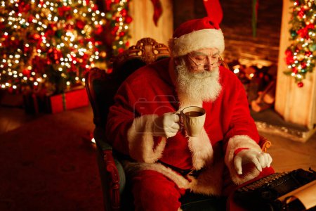 Foto de Retrato de ángulo alto de Santa Claus tradicional usando máquina de escribir y disfrutando de choco caliente en la víspera de Navidad, espacio de copia - Imagen libre de derechos