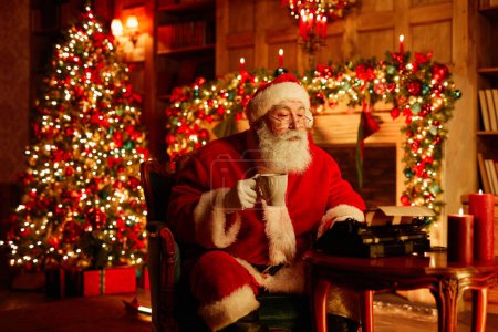 Téléchargez les photos : Portrait du Père Noël traditionnel utilisant une machine à écrire et appréciant le chocolat chaud la veille de Noël - en image libre de droit