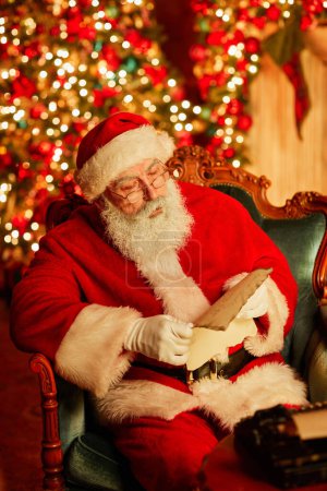 Photo for Vertical portrait of traditional Santa Claus reading letter by fireplace on Christmas eve - Royalty Free Image