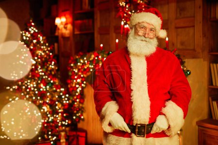 Foto de Retrato de la cintura hacia arriba de Santa Claus tradicional mirando a la cámara mientras está de pie en la habitación con árbol de Navidad y luces centelleantes, espacio de copia - Imagen libre de derechos