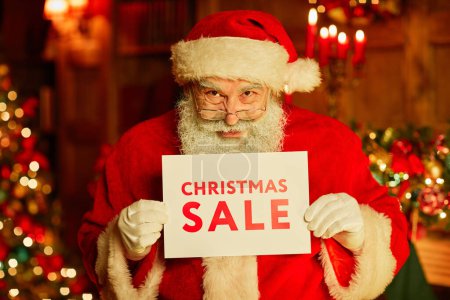 Photo for Waist up portrait of traditional Santa Claus holding Christmas SALE sign while standing in decorated room - Royalty Free Image