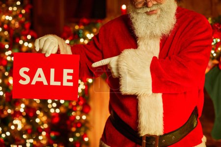 Photo for Cropped shot of traditional Santa Claus holding red SALE sign and pointing while standing in decorated room on Christmas - Royalty Free Image
