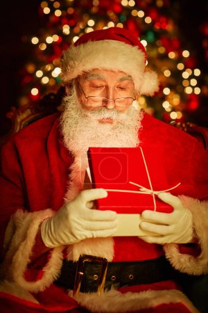 Photo for Vertical portrait of bearded traditional Santa Claus opening present with light inside by Christmas tree - Royalty Free Image