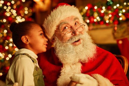 Foto de Retrato de linda chica negra susurrando secretos a Santa Claus en la víspera de Navidad - Imagen libre de derechos