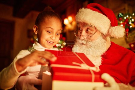 Foto de Retrato de linda niña abriendo regalo de Navidad con luz mágica mientras está sentada en el regazo de Santas - Imagen libre de derechos