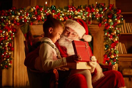 Foto de Retrato de vista lateral de una linda chica afroamericana abriendo regalo de Navidad mientras está sentada en el regazo de Santas - Imagen libre de derechos