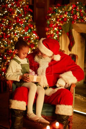Photo for Vertical portrait of cute African American girl sitting in Santas lap and holding Christmas present in cozy setting - Royalty Free Image