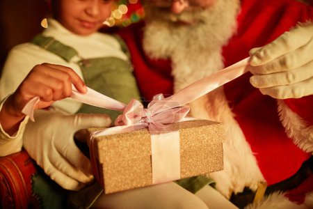 Photo for Close up of little girl opening Christmas present while sitting in Santas lap, copy space - Royalty Free Image