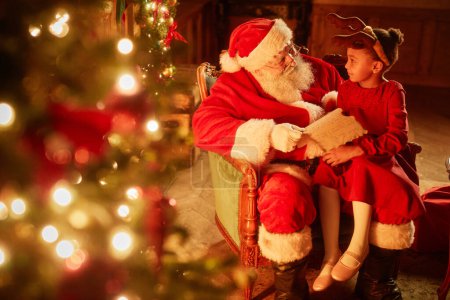 Photo for Portrait of cute little girl sitting in Santas lap by Christmas tree in magical set, copy space - Royalty Free Image