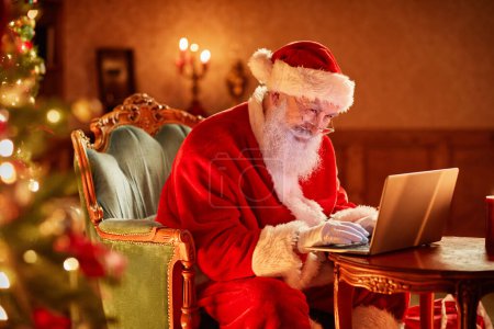 Téléchargez les photos : Portrait latéral du Père Noël à l'aide d'un ordinateur portable près du sapin de Noël et souriant, espace de copie - en image libre de droit