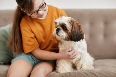 Foto de Retrato de linda chica con síndrome de Down jugando con un perro pequeño mientras están sentados en el sofá juntos, espacio de copia - Imagen libre de derechos