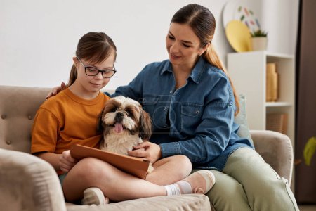Photo for Portrait of loving mother and daughter with Down syndrome playing with dog while sitting on couch at home - Royalty Free Image