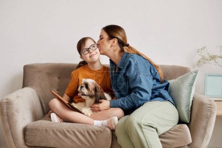 Téléchargez les photos : Portrait minimal de mère attentionnée embrassant sa fille avec le syndrome de Down sur la joue alors qu'elle était assise ensemble sur le canapé à la maison - en image libre de droit