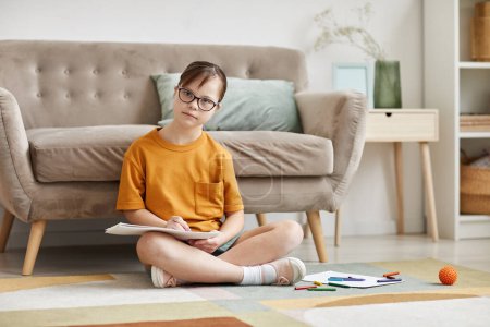 Photo for Full length portrait of teenage girl with Down syndrome sitting on floor at home cross legged and looking at camera, copy space - Royalty Free Image