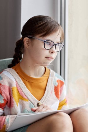 Téléchargez les photos : Portrait vertical d'une adolescente trisomique dessinant des images alors qu'elle était assise par la fenêtre à la maison - en image libre de droit