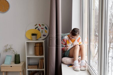 Foto de Retrato de longitud completa de la chica adolescente dibujando imágenes mientras está sentado junto a la ventana en una habitación acogedora, espacio de copia - Imagen libre de derechos