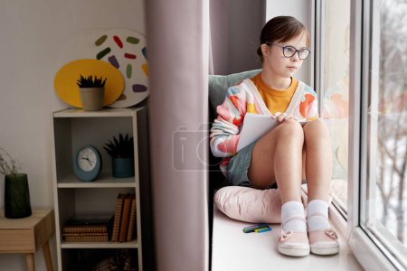 Photo for Full length portrait of teenage girl with Down syndrome drawing pictures while sitting by window in cozy room, copy space - Royalty Free Image