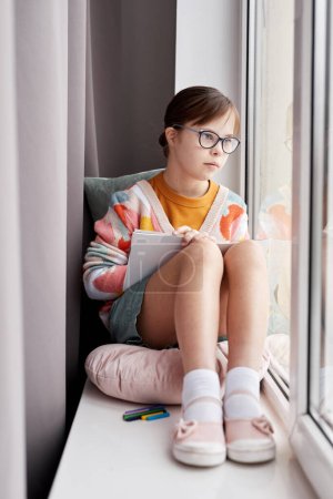Photo for Vertical portrait of teenage girl with Down syndrome drawing pictures while sitting by window in cozy room - Royalty Free Image