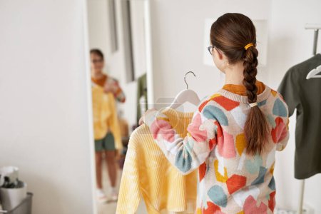 Photo for Back view at teenage girl with Down syndrome choosing clothes and looking at mirror at home, copy space - Royalty Free Image