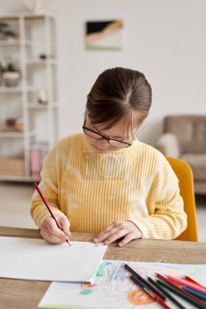 Photo for Vertical portrait of teenage girl with Down syndrome drawing pictures at table in cozy room - Royalty Free Image