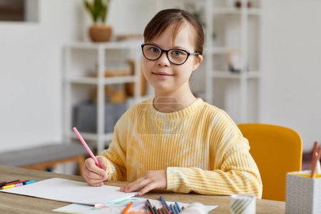 Photo for Portrait of cute teen girl with Down syndrome drawing pictures while sitting at desk and smiling at camera - Royalty Free Image
