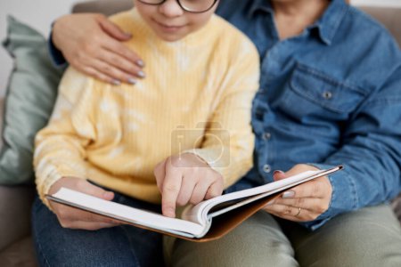 Photo for Close up of young girl with Down syndrome reading book with loving mother at home - Royalty Free Image