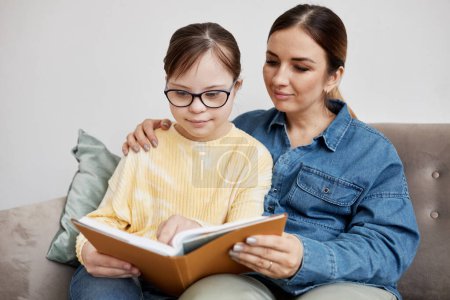 Photo for Portrait of teen girl with Down syndrome reading book with loving mother at home - Royalty Free Image