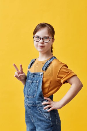 Photo for Vertical portrait of teen girl with Down syndrome looking at camera and showing peace sign while standing against yellow background in studio - Royalty Free Image