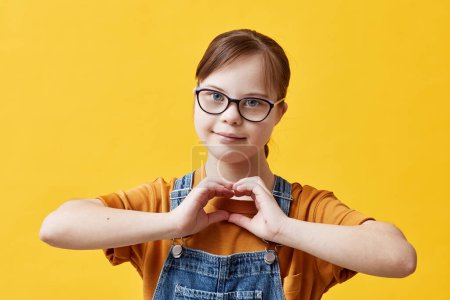 Photo for Waist up portrait of teen girl with Down syndrome looking at camera and showing heart sign while standing against yellow background in studio - Royalty Free Image