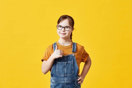 Photo for Portrait of cute girl with Down syndrome looking at camera against yellow background in studio and showing thumbs up - Royalty Free Image