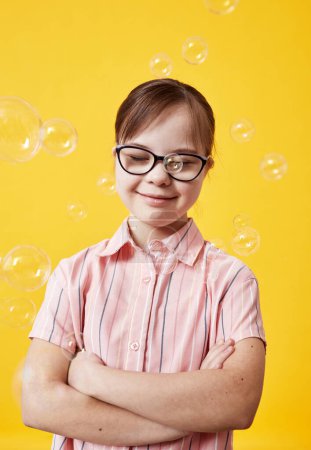 Photo for Vertical portrait pf playful cute girl with Down syndrome playing with bubbles against yellow background - Royalty Free Image