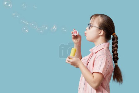 Photo for Side view portrait pf playful girl with Down syndrome blowing bubbles against blue background - Royalty Free Image