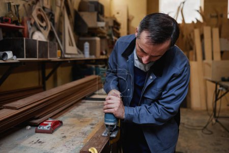 Téléchargez les photos : Portrait d'un ouvrier masculin mature découpant du bois dans un atelier d'usine avec outil électrique, espace de copie - en image libre de droit