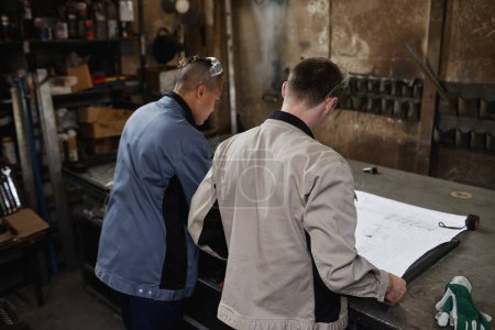 Photo for Back view of two workers looking at blueprints and plans in industrial factory workshop - Royalty Free Image