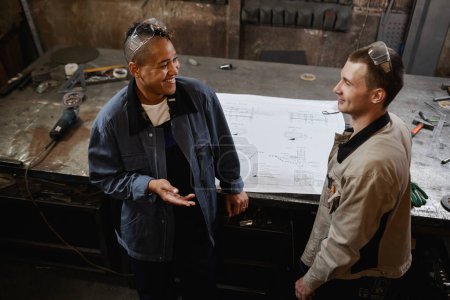 Foto de Retrato de alto ángulo de dos trabajadores de la fábrica sonrientes discutiendo proyecto mientras están de pie en el taller - Imagen libre de derechos
