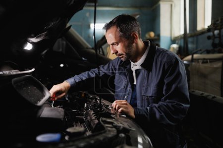 Téléchargez les photos : Portrait latéral du moteur de voiture de fixation de mécanicien mature dans le garage sombre magasin - en image libre de droit