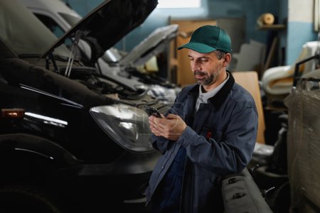 Foto de Retrato de cintura hacia arriba de un trabajador varón maduro usando un teléfono inteligente en un taller de fábrica de automóviles - Imagen libre de derechos
