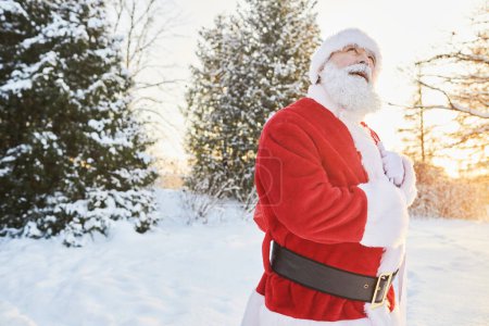 Téléchargez les photos : Portrait taille haute du Père Noël traditionnel portant un sac avec des cadeaux à l'extérieur dans la forêt d'hiver et levant les yeux, copier l'espace - en image libre de droit