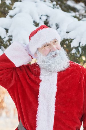 Foto de Retrato vertical de la cintura hacia arriba de Papá Noel tradicional sonriendo al aire libre en el bosque de invierno y mirando hacia otro lado - Imagen libre de derechos