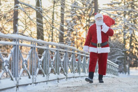 Foto de Retrato de cuerpo entero de Papá Noel tradicional llevando saco con regalos y caminando hacia la cámara en el paisaje de invierno, espacio de copia - Imagen libre de derechos