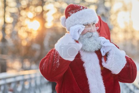 Téléchargez les photos : Portrait taille haute du Père Noël traditionnel portant un sac avec des cadeaux en plein air en hiver et regardant au-dessus des lunettes, espace de copie - en image libre de droit