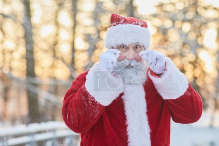 Photo for Waist up portrait of traditional Santa Claus looking at camera over eyeglasses and looking surprised in winter forest outdoors, copy space - Royalty Free Image