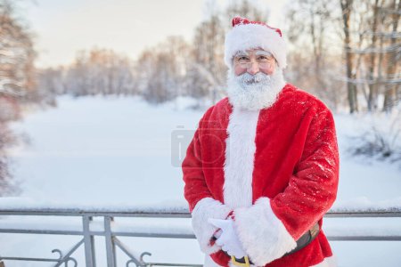 Téléchargez les photos : Portrait taille haute du Père Noël traditionnel regardant la caméra et souriant dans le parc d'hiver, espace de copie - en image libre de droit