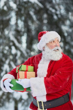 Foto de Cintura hacia arriba retrato de Santa Claus tradicional llevando regalos al aire libre en el bosque de invierno y buscando cauteloso - Imagen libre de derechos