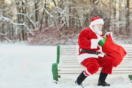 Photo for Side view portrait of traditional santa Claus holding bag with presents while sitting on bench outdoors, copy space - Royalty Free Image