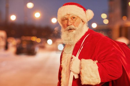 Foto de Retrato de la cintura hacia arriba de Santa Claus tradicional caminando en la ciudad por la noche y mirando a su alrededor, espacio de copia - Imagen libre de derechos