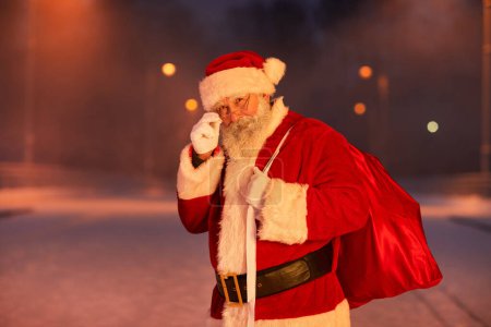 Téléchargez les photos : Portrait taille haute du Père Noël traditionnel debout dans la rue de la ville la nuit et regardant la caméra la veille de Noël, copier l'espace - en image libre de droit