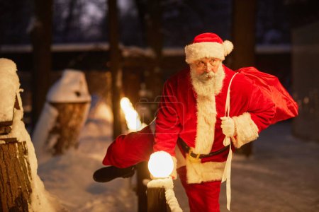 Foto de Retrato de Papá Noel tradicional con saco de regalos trepando por la valla al aire libre en la víspera de Navidad - Imagen libre de derechos
