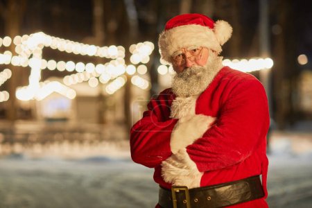 Photo for Waist up portrait of Santa Claus looking at camera while standing outdoors at night with arms crossed, copy space - Royalty Free Image