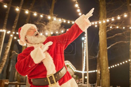 Téléchargez les photos : Portrait taille haute du Père Noël dansant à l'extérieur la nuit et soufflant de la fumée, copiant l'espace - en image libre de droit